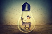 Woman working on laptop computer sitting inside electric light bulb isolated on gray office wall background