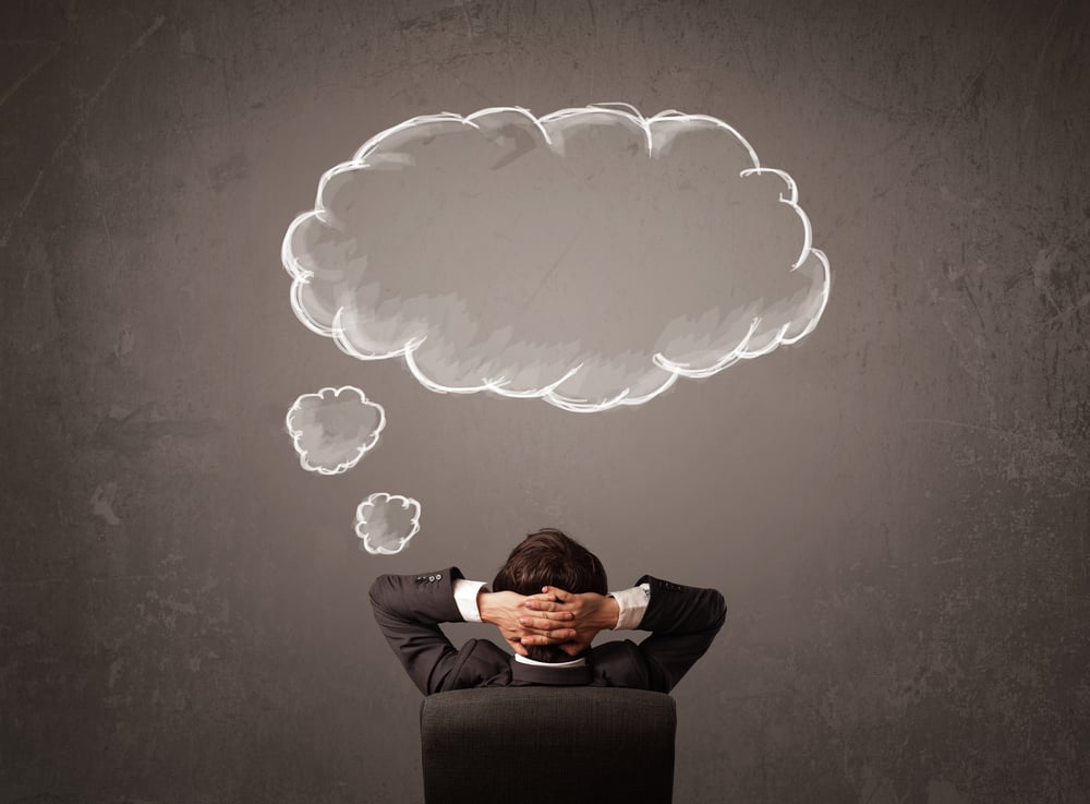 Young businessman sitting in office chair in front of a wall with cloud thought sketched on a chalkboard above his head-1